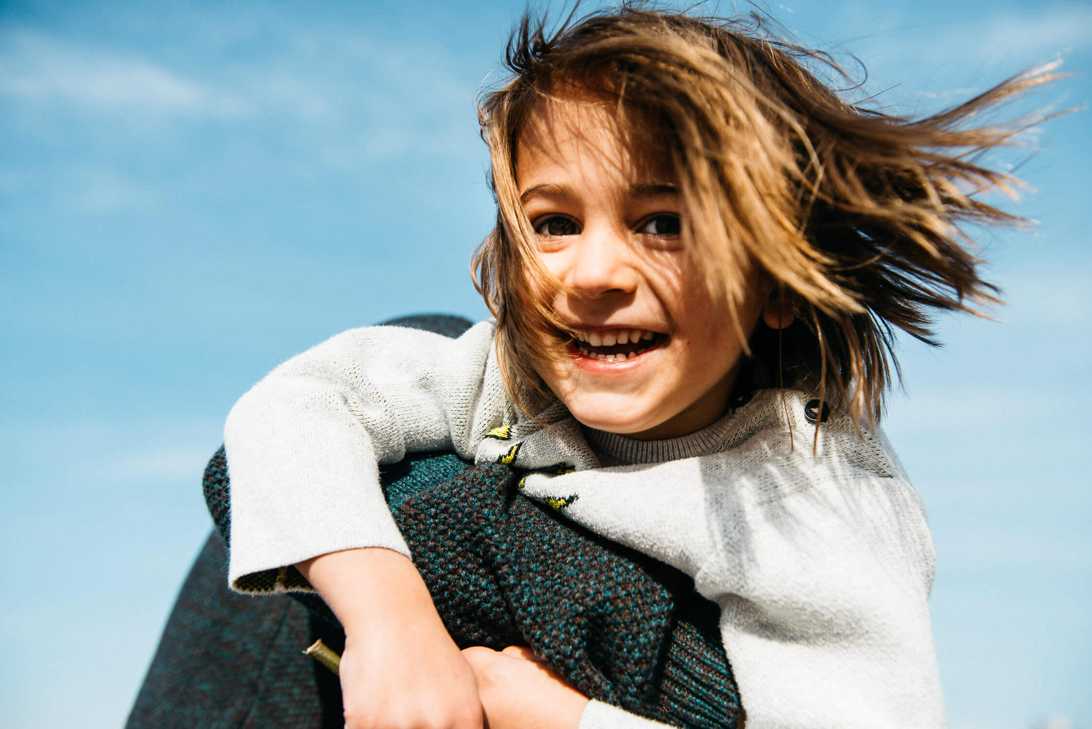 A smiling child with windblown hair embraces someone against a backdrop of a clear blue sky. The child wears a light-colored sweater and appears joyful and carefree.