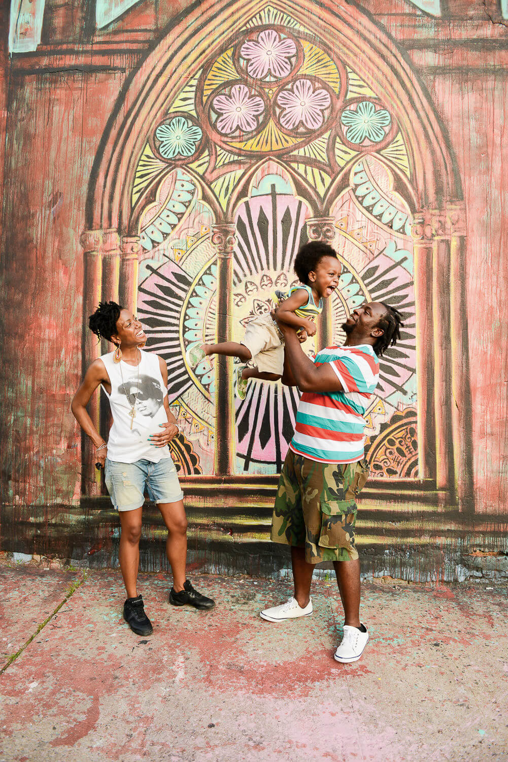 A joyful family scene in front of a colorful mural depicting a large, decorative arched window. One person playfully lifts a child in the air, while another watches and smiles. The background features vibrant patterns and details.