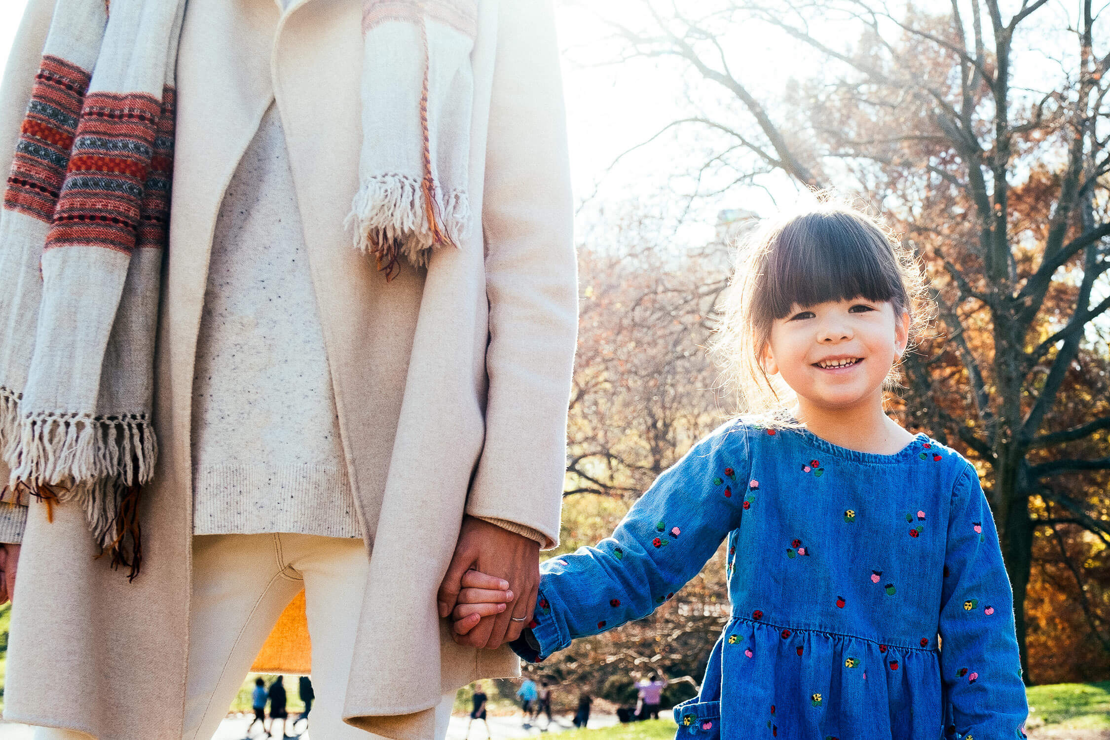 A young girl in a blue dress holds hands with an adult wearing a beige coat and scarf. They are outdoors in a park on a sunny day with trees and people in the background. The girl is smiling at the camera.