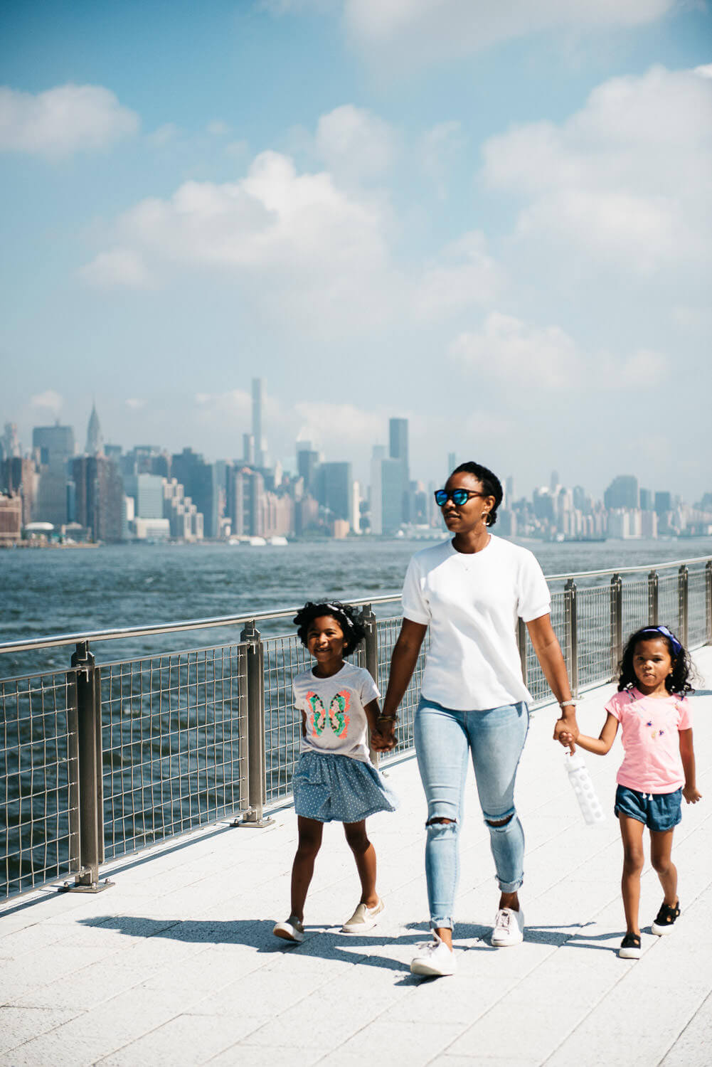 A woman walks with two children along a waterfront promenade on a sunny day. The background features a city skyline with skyscrapers and a blue sky with clouds. The woman wears sunglasses, a white t-shirt, and jeans. The children wear colorful outfits.
