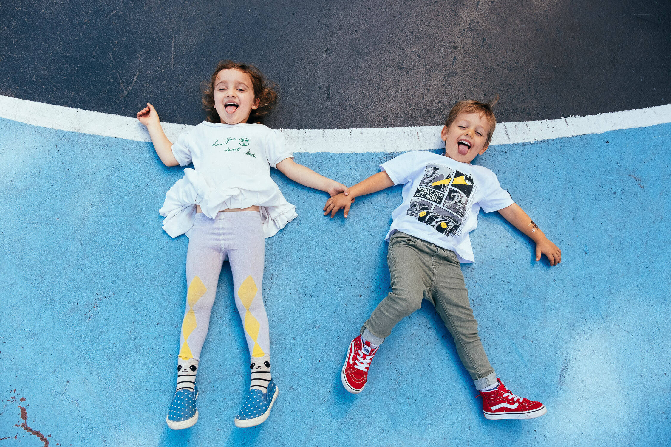 two children laughing while lying on a blue playground surface, holding hands