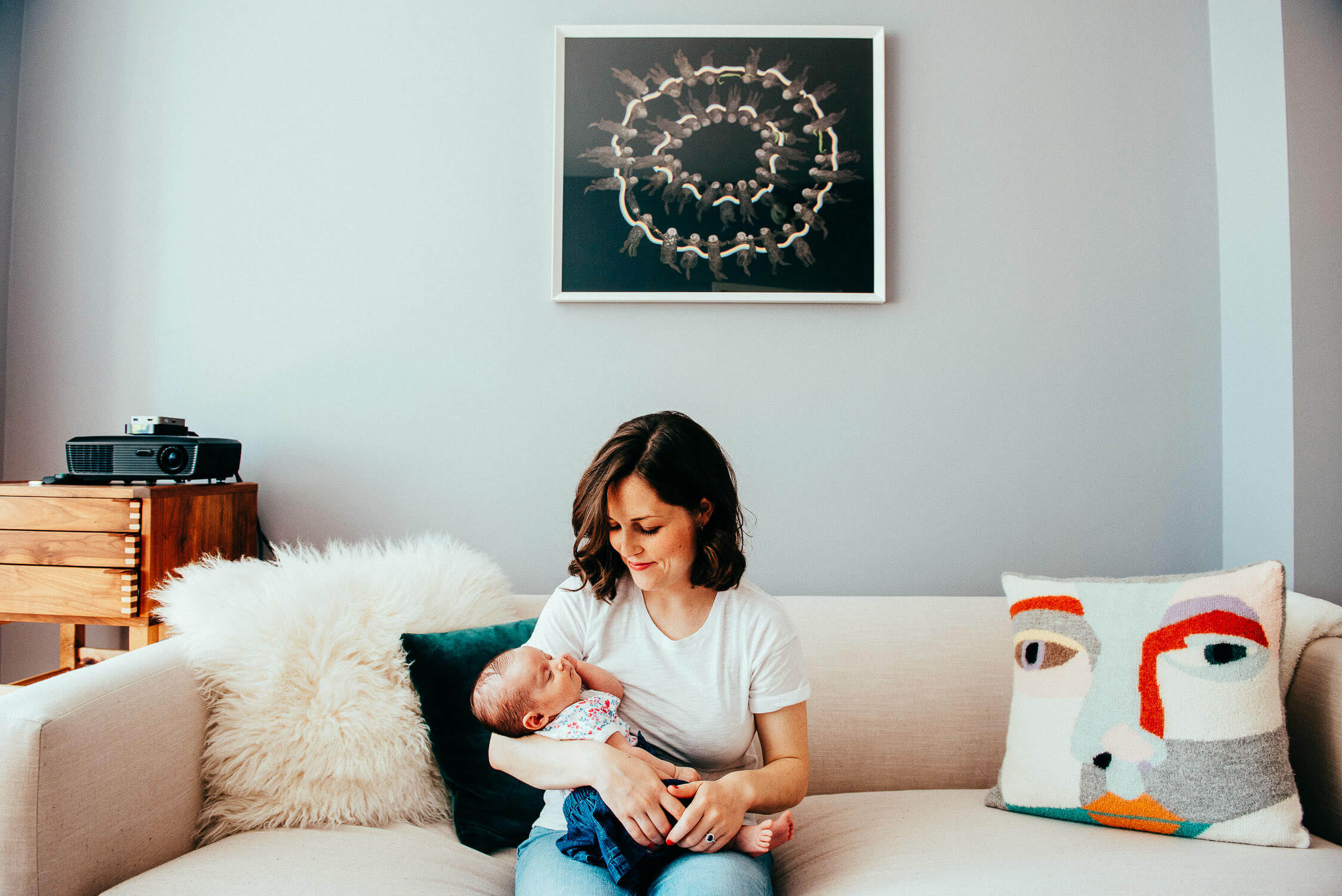 A woman sitting on a couch holds a baby in her arms. The room features a modern decor with a unique art piece on the wall and a colorful abstract pillow. A projector sits on a side table with a fluffy cushion beside her.