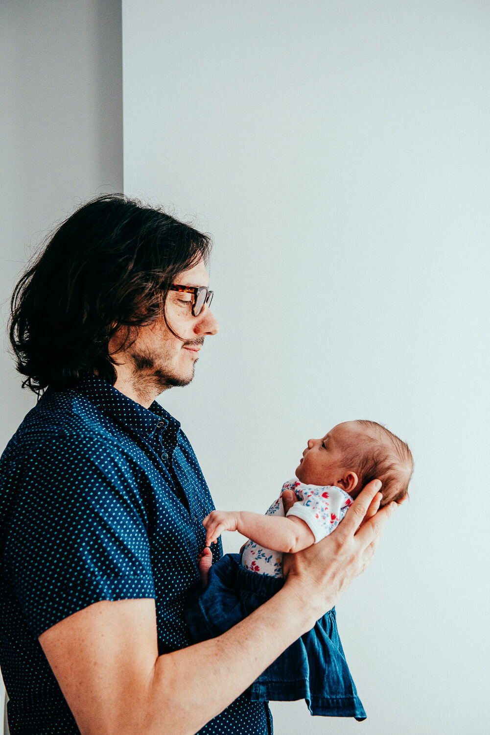 A person with long dark hair and glasses is holding a baby. The baby is wearing a colorful outfit. They are standing against a plain background, gazing at each other.