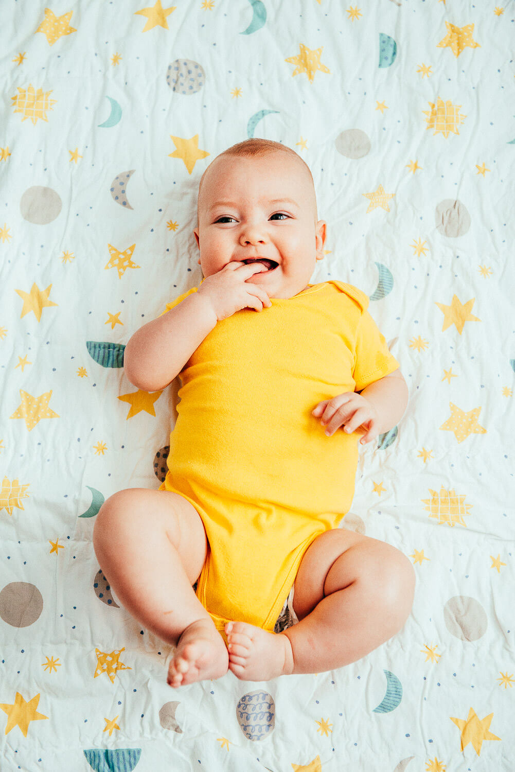 A smiling baby in a yellow onesie lies on a patterned blanket with moons, stars, and clouds. The baby is playfully touching their mouth with one hand.