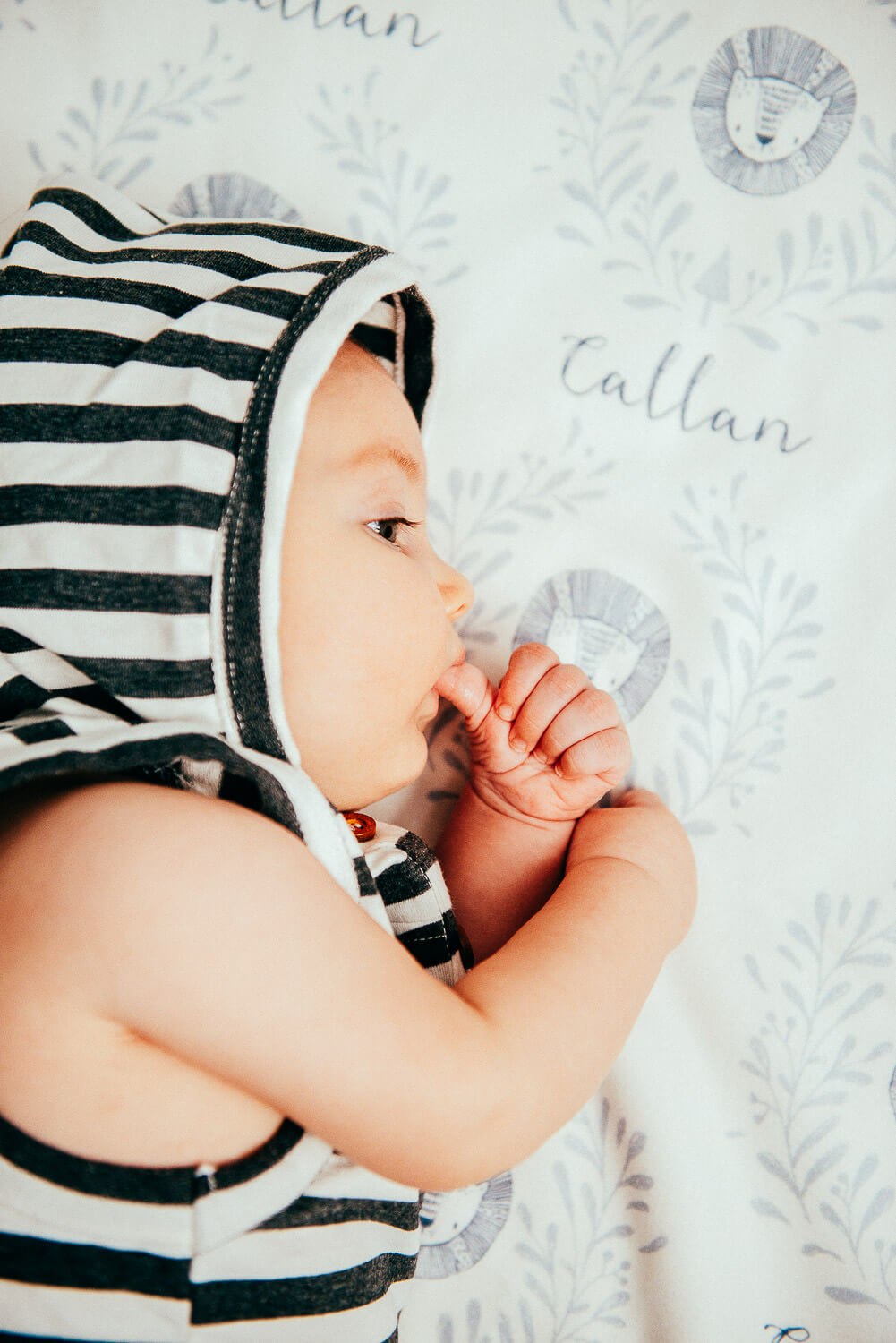 A baby wearing a striped hooded outfit lies on a personalized blanket with the name Callan and lion illustrations. The baby is sucking their thumb and is facing sideways.