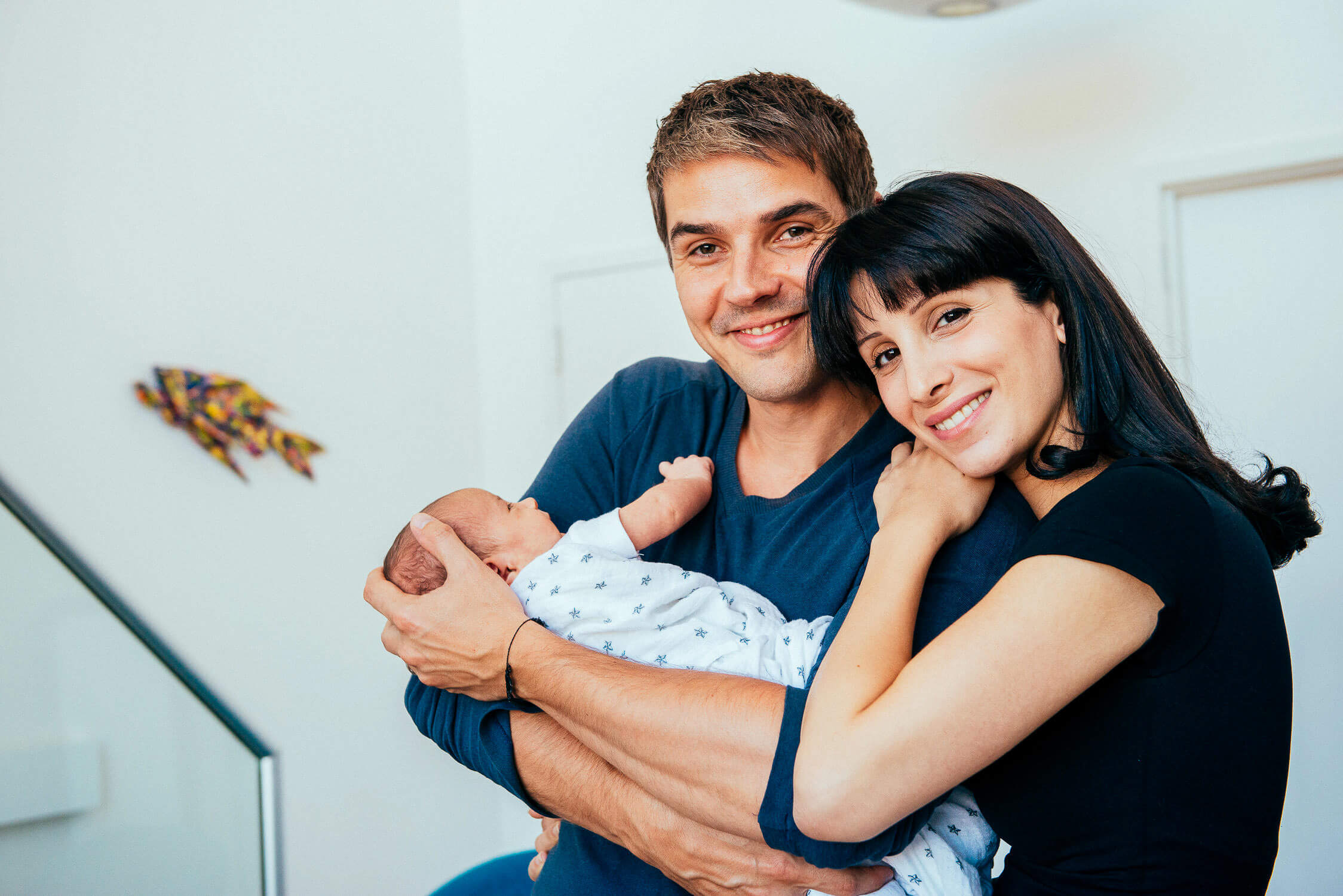 A couple smiling and holding a newborn baby wrapped in a patterned blanket. They stand in a brightly lit room with modern decor visible in the background.