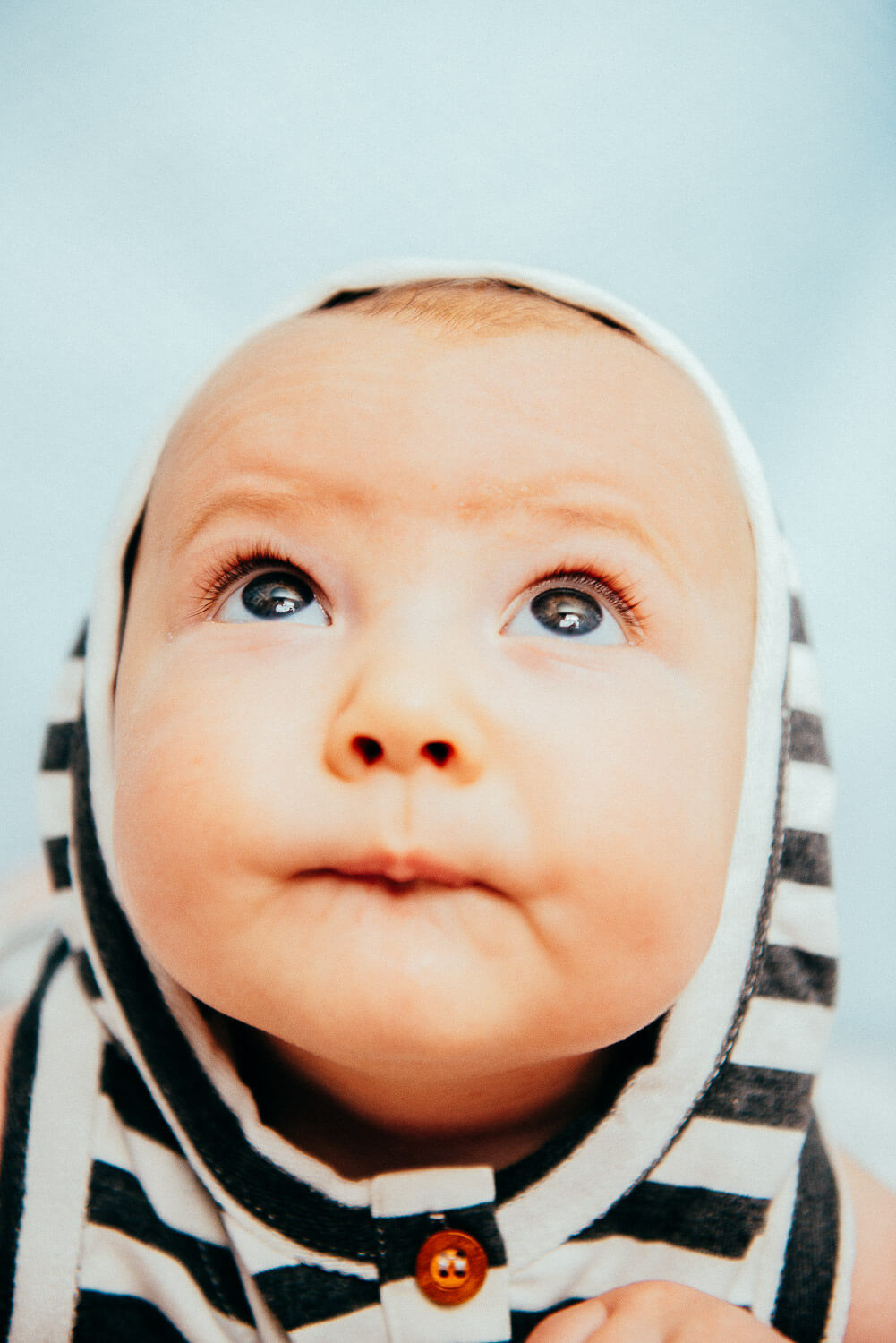 A baby wearing a striped hooded outfit looks upwards with wide eyes. The hood has black and white stripes, and the background is a soft blue. The baby has a curious expression.