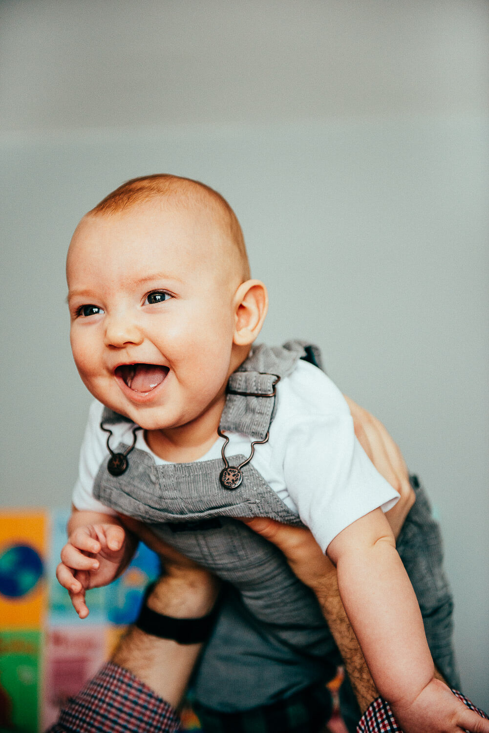 A smiling baby in gray overalls and a white shirt is being held up playfully by an adult. The background is blurred with colorful elements.