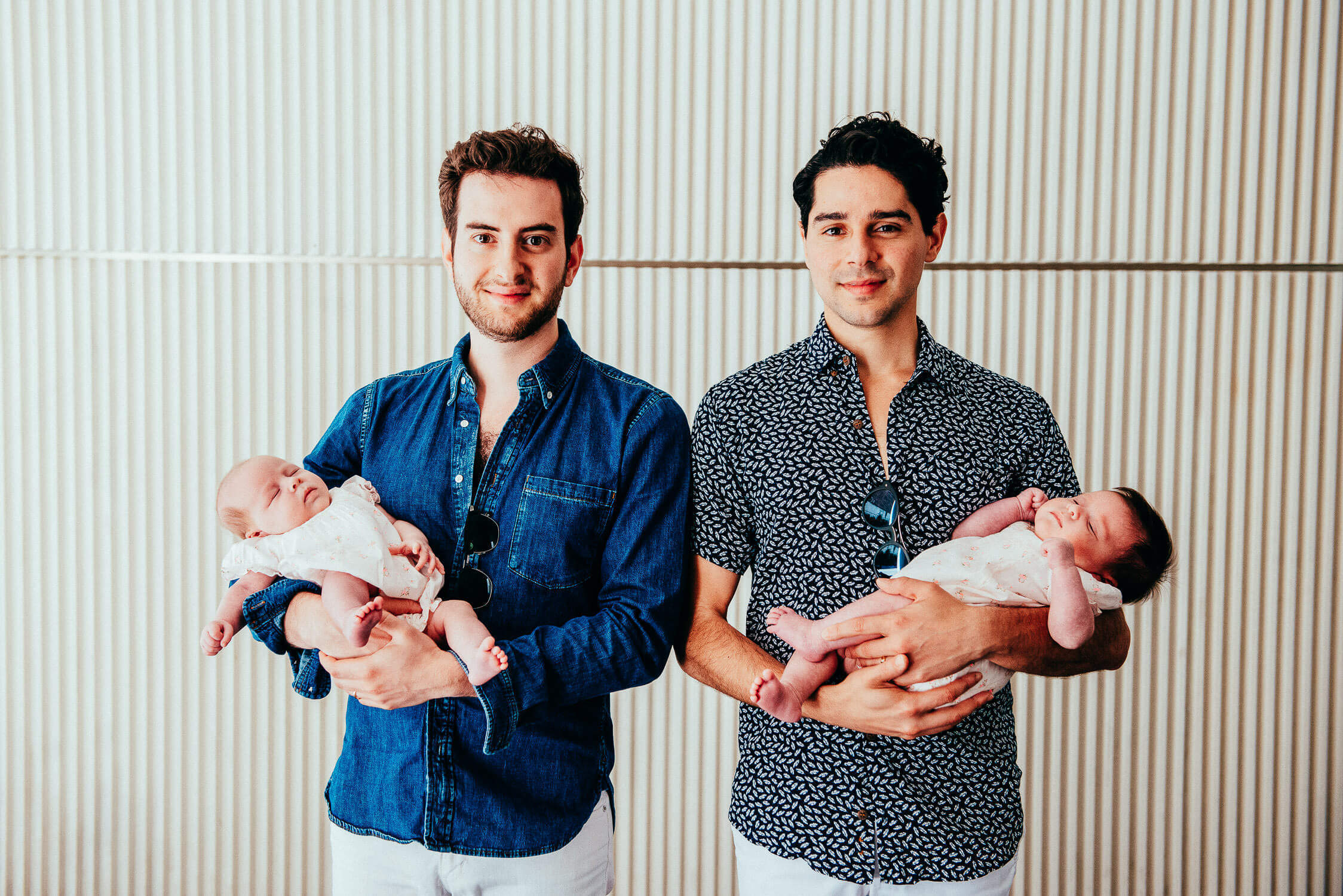 Two men are standing side by side, each holding a baby. The man on the left wears a denim shirt, while the man on the right wears a patterned shirt. Both babies are dressed in light-colored outfits and are sleeping peacefully. The background is striped.