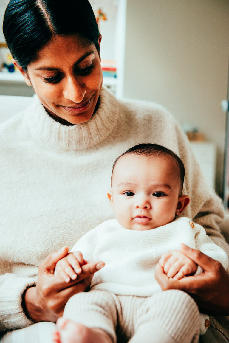 A person wearing a cream sweater gently holds a baby dressed in a matching outfit, sitting on their lap. The person looks down at the baby with a warm smile in a cozy indoor setting.