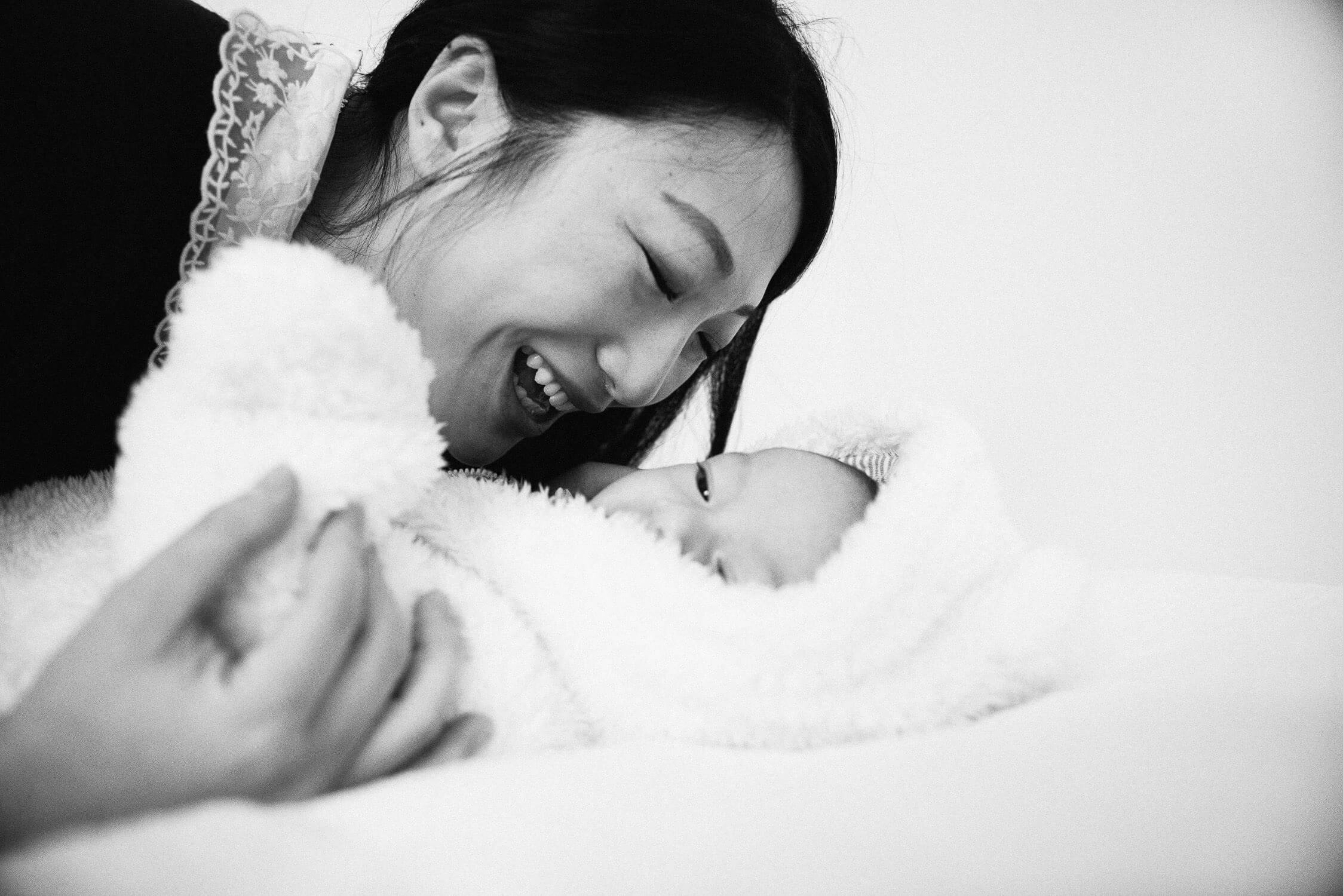 A black and white image shows a woman gently smiling down at a baby dressed in a fluffy outfit with bear ears. They are lying on a soft surface, creating a tender and joyful moment.