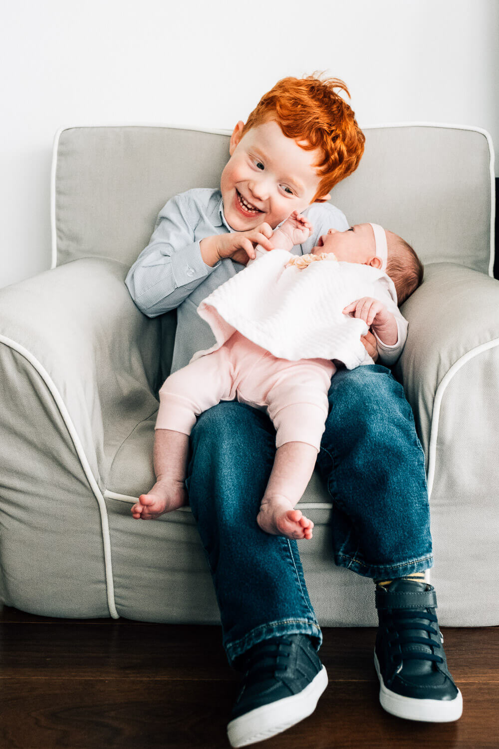 A smiling young boy with red hair sits on a gray armchair, holding a sleeping baby wearing a white and pink outfit. The boy is gently touching the baby’s face.