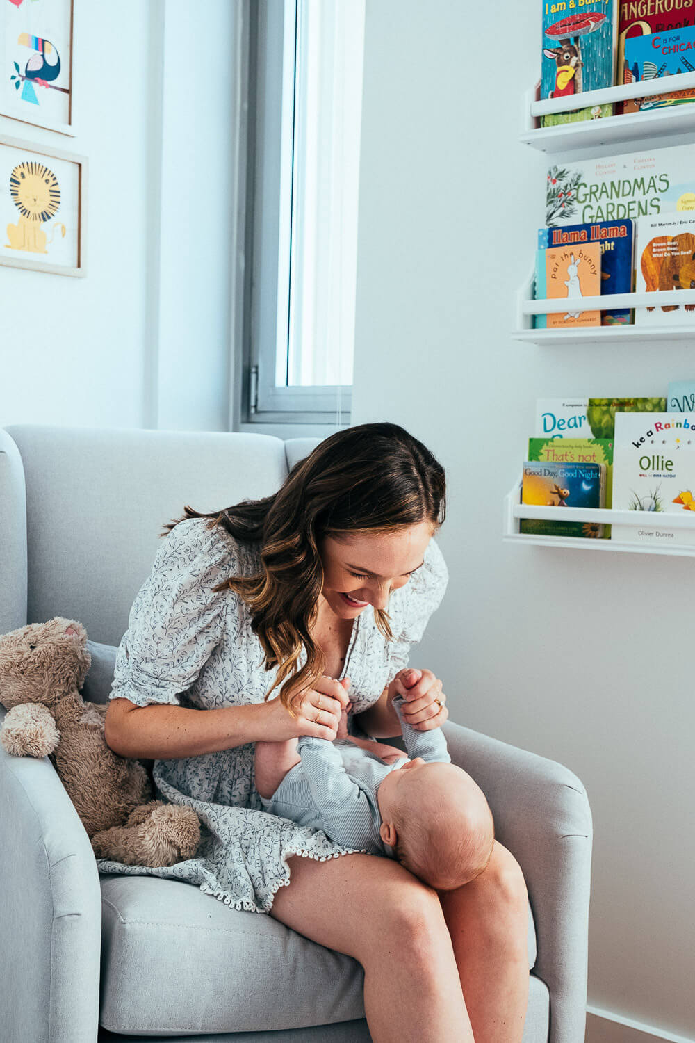 A woman sits in a light gray armchair, smiling at a baby on her lap. A plush teddy bear is beside her. A wall with childrens books on shelves is in the background, and a window lets in natural light.