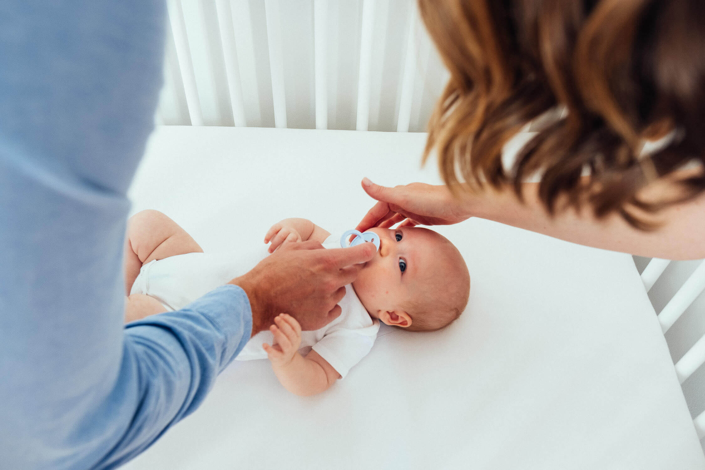 Two adults gently tend to a baby lying in a crib. The baby is wearing a white onesie and holding a pacifier. The adults hands are visible as they adjust the babys position, creating a nurturing and caring scene.