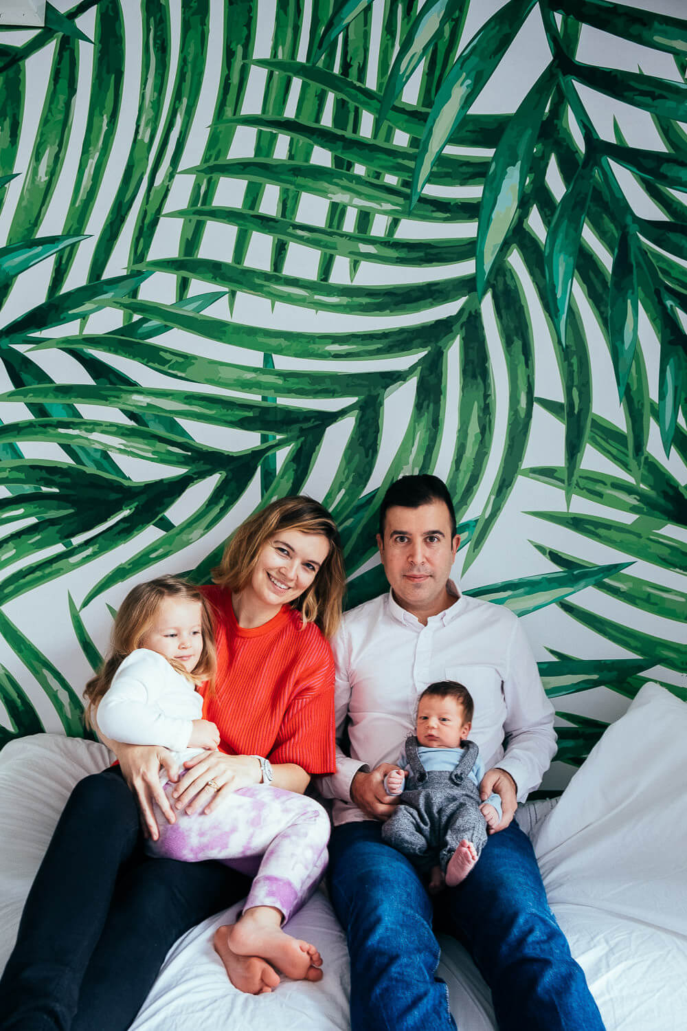A family of four sits on a white couch in front of a leafy green wallpaper. The woman in a red sweater holds a young child, while the man in a white shirt holds a baby. They are all smiling warmly at the camera.