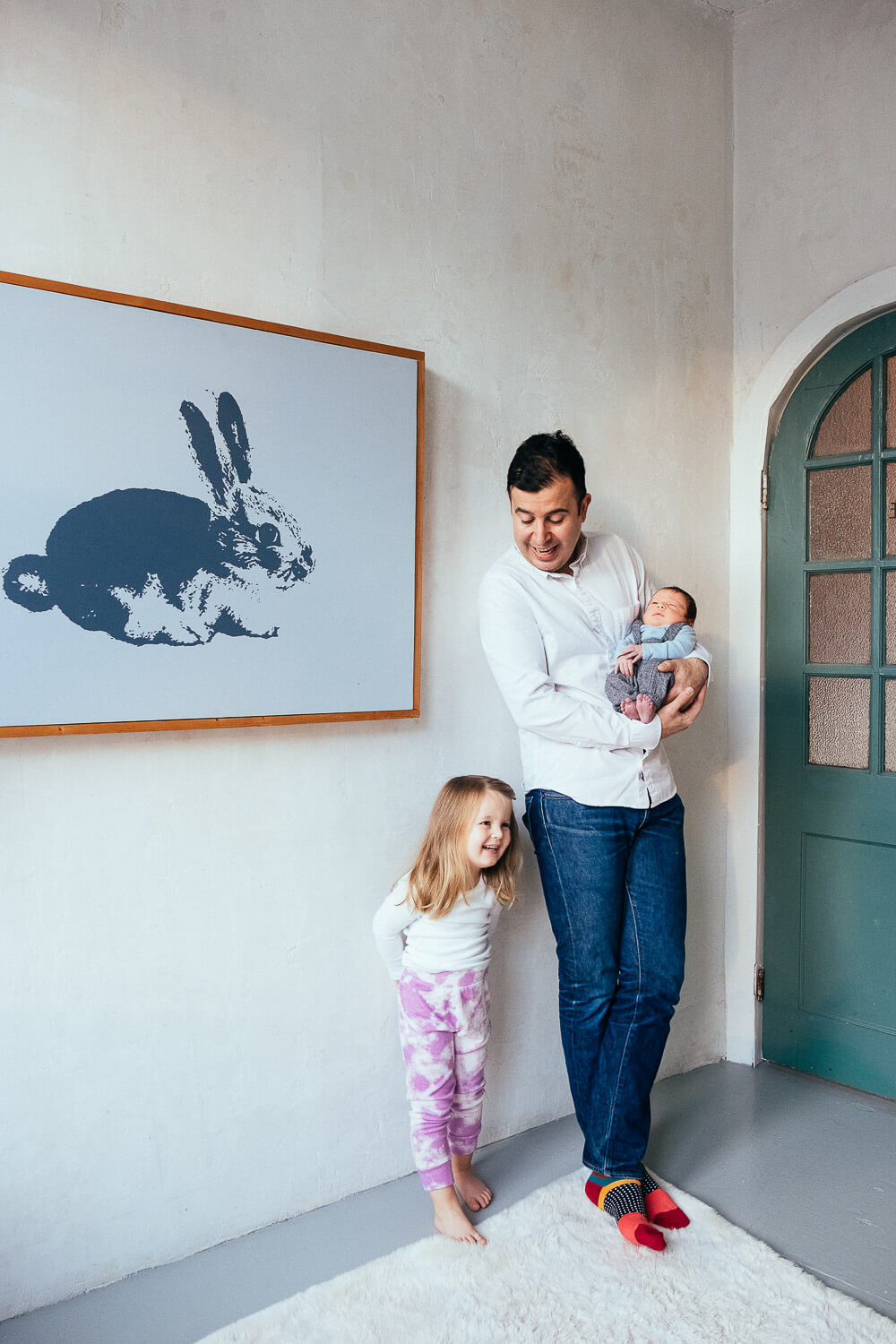 Father standing next to a young daughter, holding a newborn baby, with a large rabbit illustration on the wall behind them.