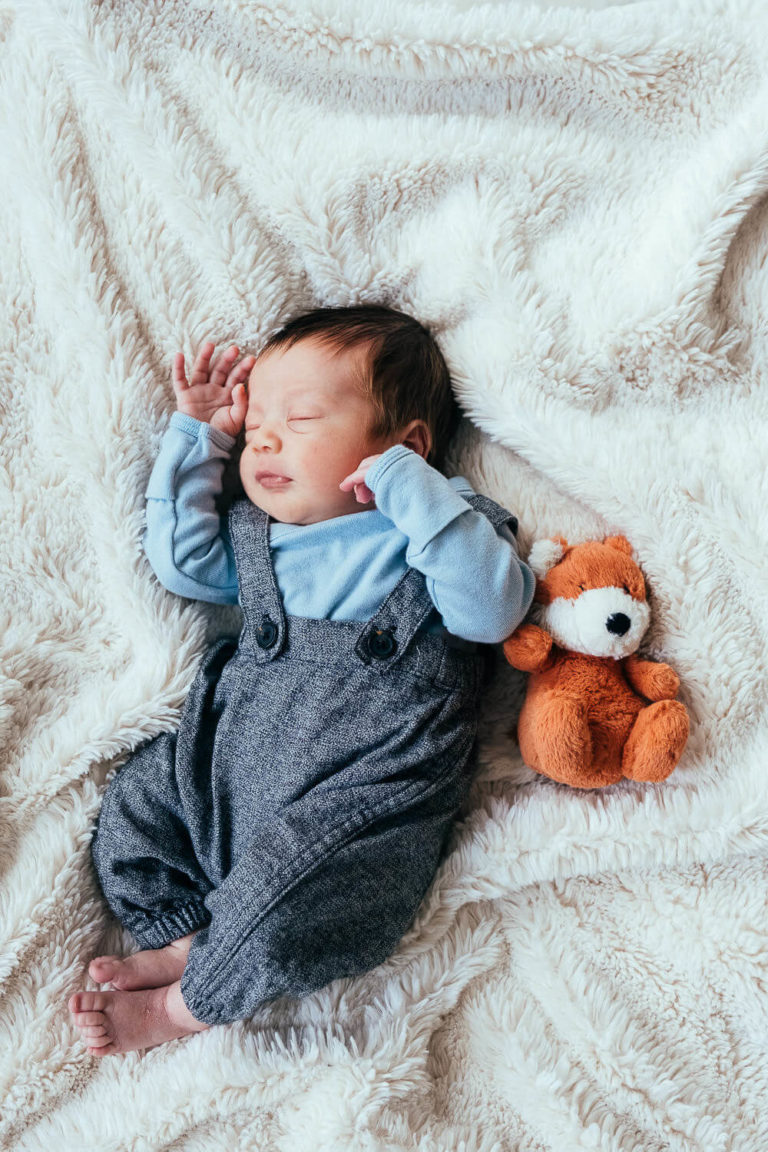 A sleeping baby in a blue shirt and gray overalls lies on a soft, white blanket. A small plush fox is beside them. The baby has dark hair and is peacefully resting with one hand near their face.
