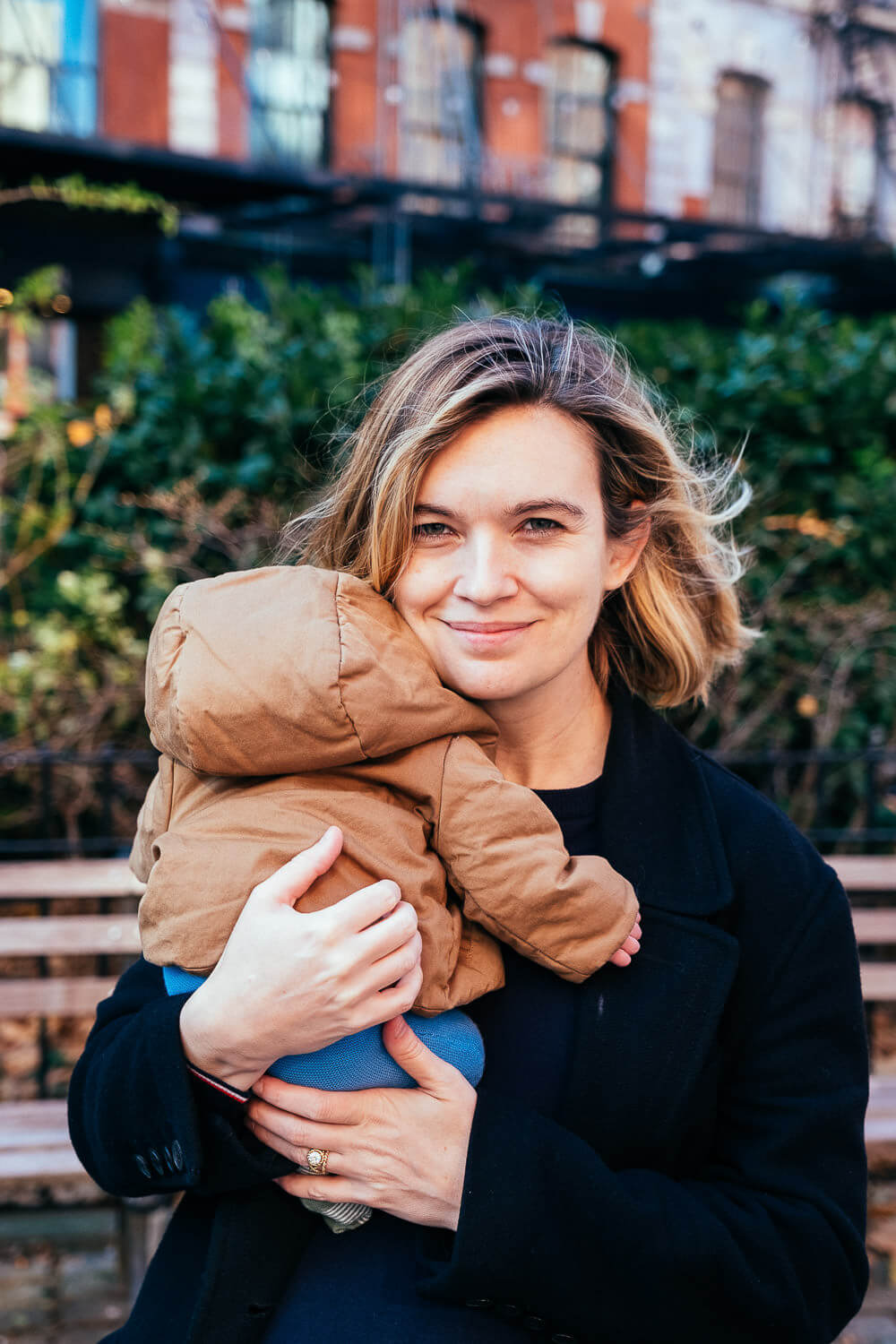 A woman is standing outdoors, smiling at the camera while holding a baby in a brown hooded jacket. They are in front of a blurred green shrubbery and brick building background. The setting appears to be a park or garden.