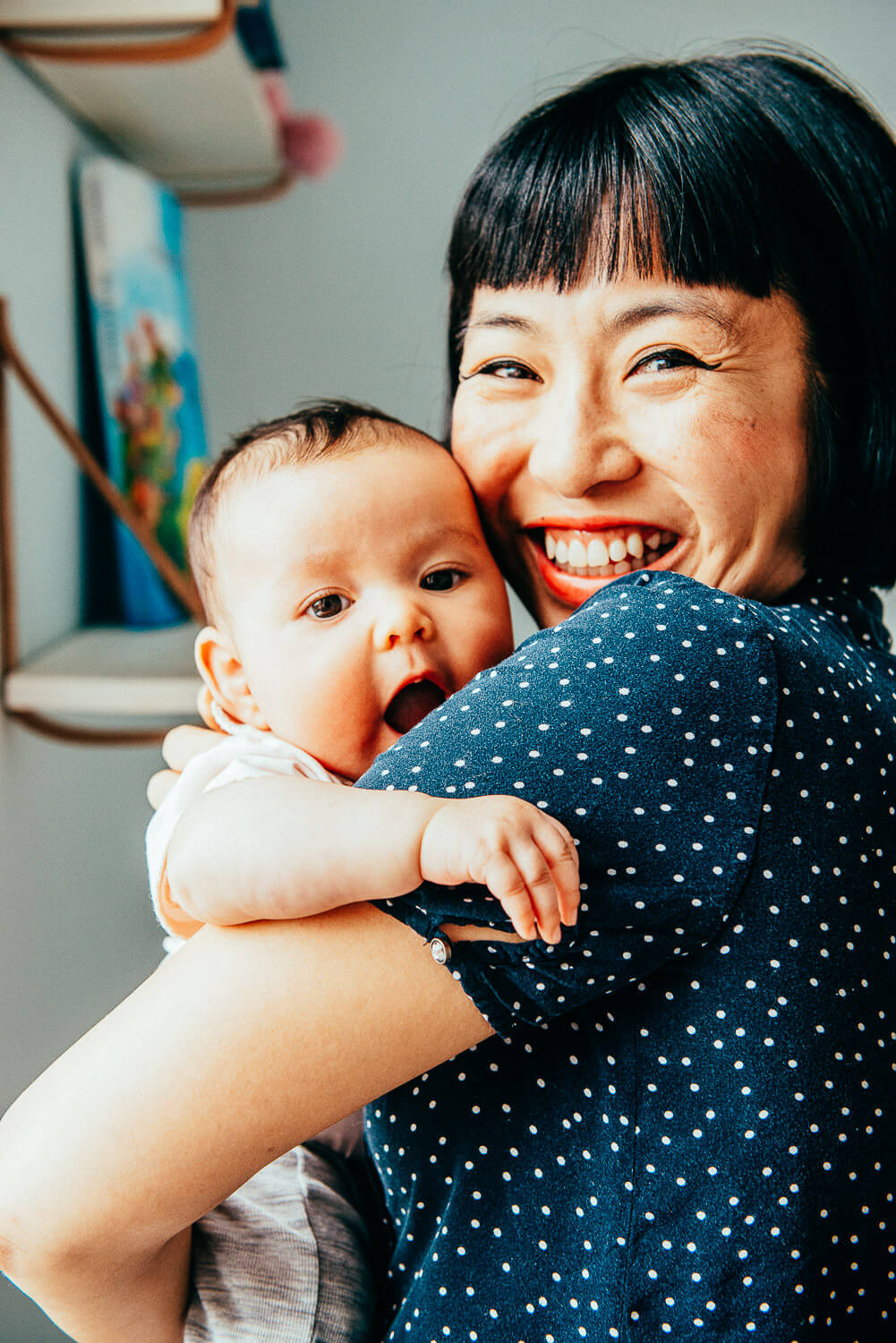 mother smiling while holding her newborn baby, who is looking over her shoulder in a bright, cozy room