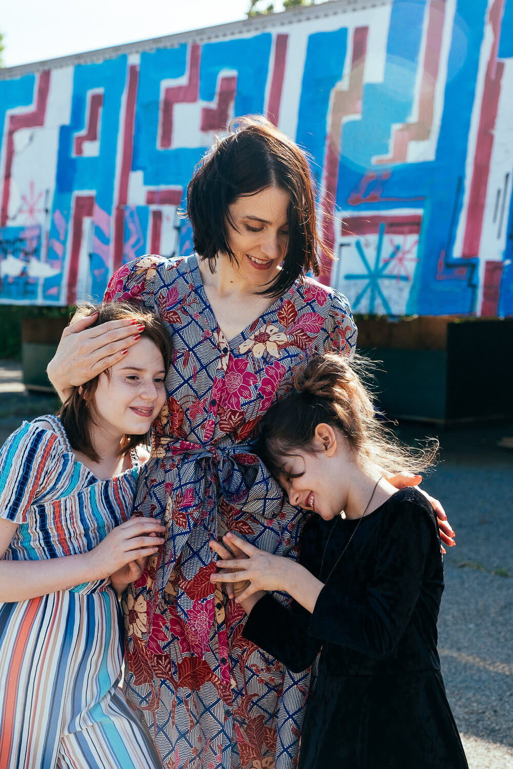 A woman in a floral dress stands with two young girls in striped and black dresses, all smiling for their Brooklyn family photographer. They are posed in front of a colorful, graffiti-covered wall that perfectly captures the vibrant essence of family photography NYC.