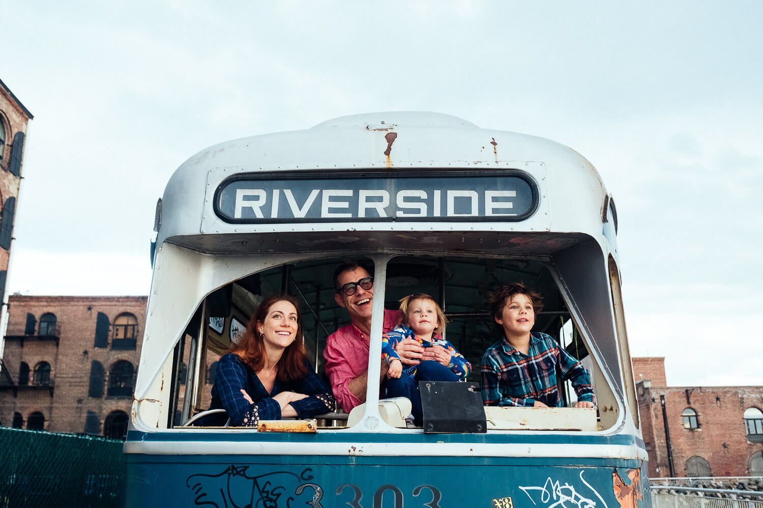 A family of four smiles inside an old bus labeled Riverside. Captured by a Brooklyn family photographer, the parents sit in the front while their children, each clutching a toy, sit beside them. The scene is set against an industrial-style building under a clear sky, showcasing family photography NYC style.
