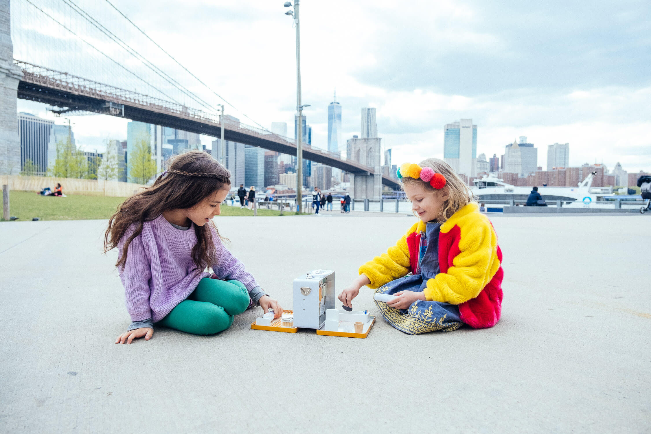 Two children, in a scene perfect for brand photography, play a tabletop game on a concrete surface in the park. One sports a purple sweater and green pants, while the other wears a colorful jacket, with the Brooklyn Bridge and city skyline painting an iconic backdrop.