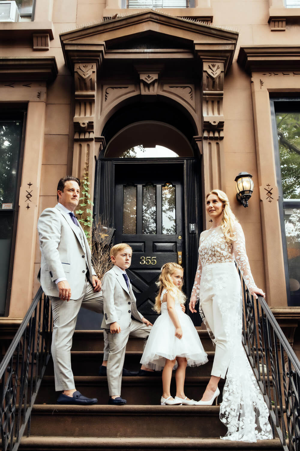 A family of four poses on the steps of a brownstone building, a classic subject for any Brooklyn family photographer. The parents wear formal attire, with the mother in a white lace gown. Their young son and daughter, also elegantly dressed, stand between them by the door numbered 355.