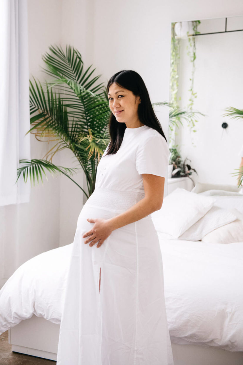 Pregnant woman in a white dress holding her belly, standing in a bright, plant-filled bedroom, capturing a serene maternity photography session in NYC.