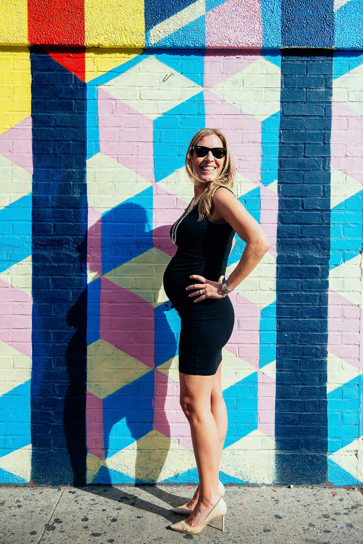 A confident and beautiful mom-to-be wearing a fitted black maternity dress, cool sunglasses, and stylish heels. Showcasing chic maternity fashion