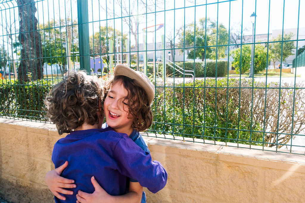 Two young children with curly hair embrace warmly during a playful photo session outdoors. One child wears a hat and smiles. They stand in front of a wire fence, with trees and a playground visible in the background, highlighting their cheerful kids friendship in the sunny setting.