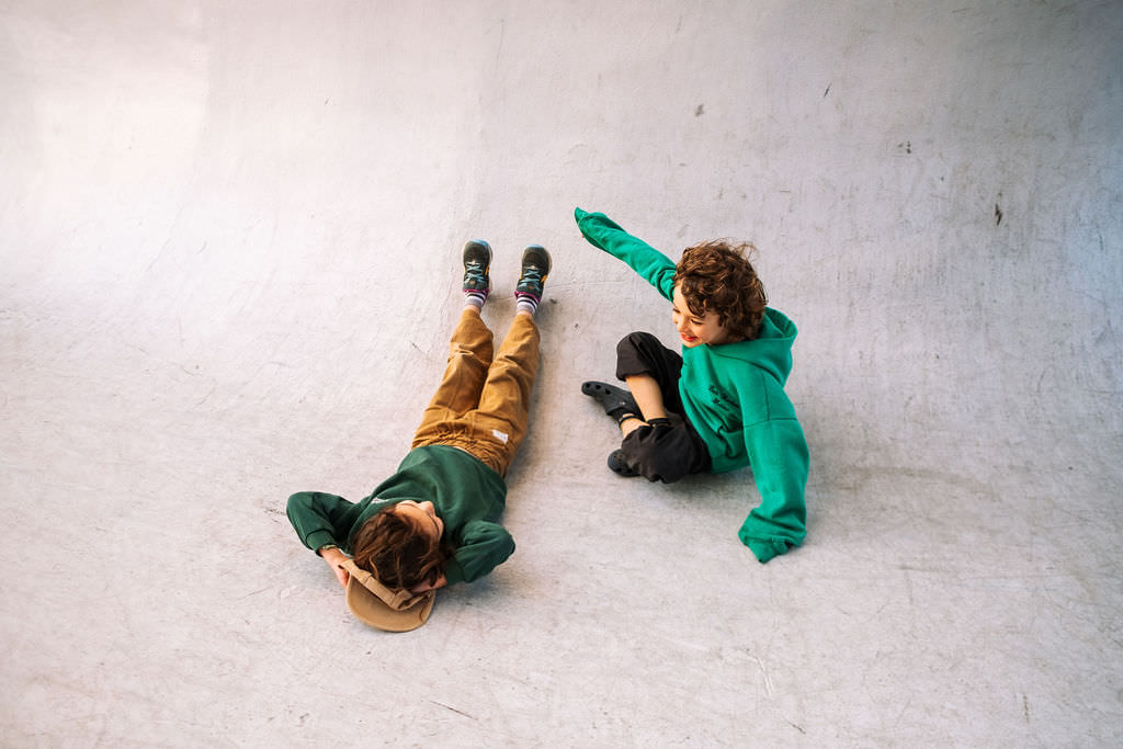 Two children celebrate their friendship on a smooth, curved concrete surface. One lies down in brown pants and a green sweater, holding a hat, while the other sits nearby in black pants and a green hoodie, capturing this playful photo session with laughter and smiles.