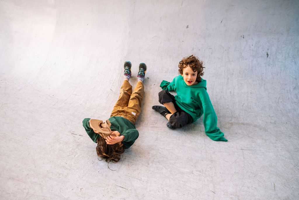 Two children are joyfully playing on a smooth, curved surface. One lies back in a green hoodie and brown pants, covering their face, while the other sits smiling at the camera in matching attire. Its a playful photo session that captures the essence of kids friendship.