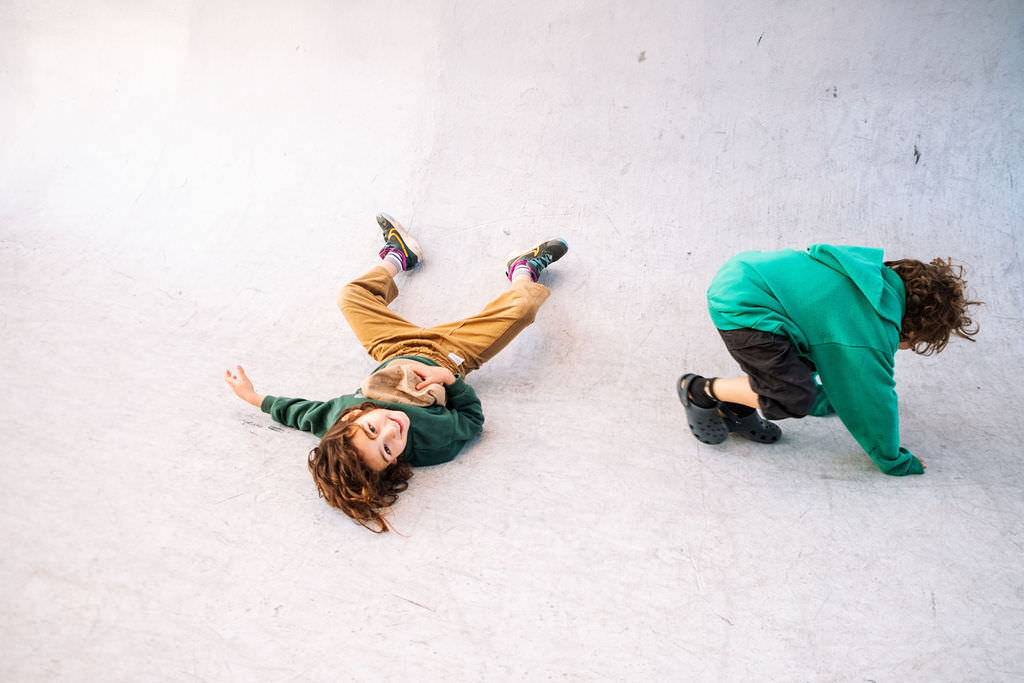 Two children with long hair engage in a playful photo session on a smooth surface. One lies back with legs up, clad in brown pants and a green hoodie, while the other crouches nearby in black shorts and a matching hoodie. Both exude the joy of kids friendship.