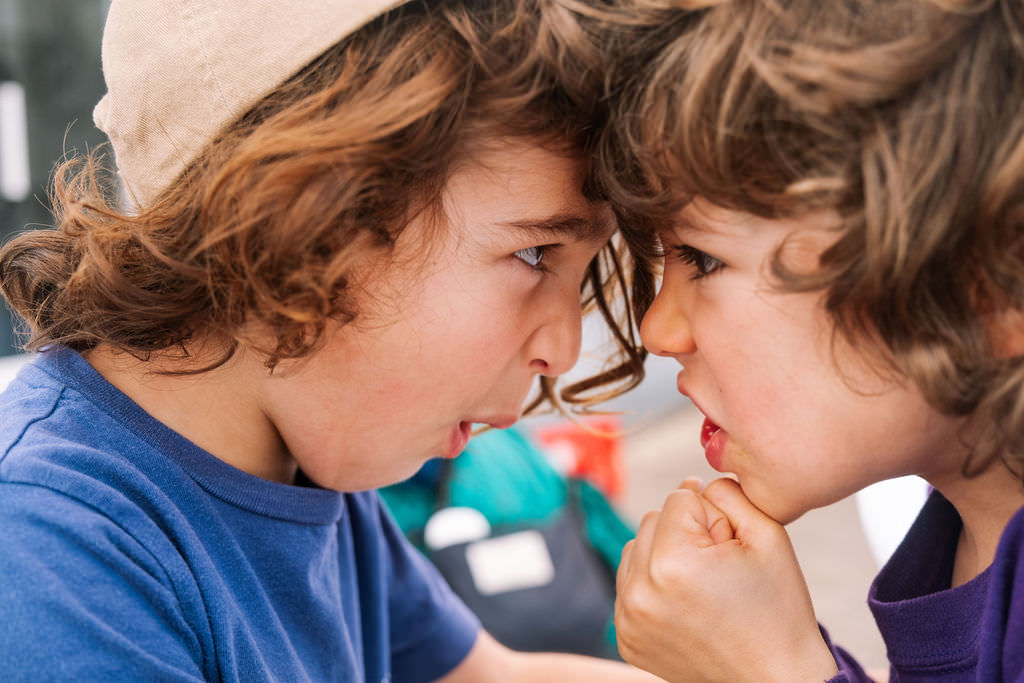 In a fun and playful photo session, two young children face each other with intense expressions. One wears a light cap, and the other sports curly hair. The child in blue appears to be in a playful confrontation, fists raised against the colorful background—a snapshot of kids friendship.