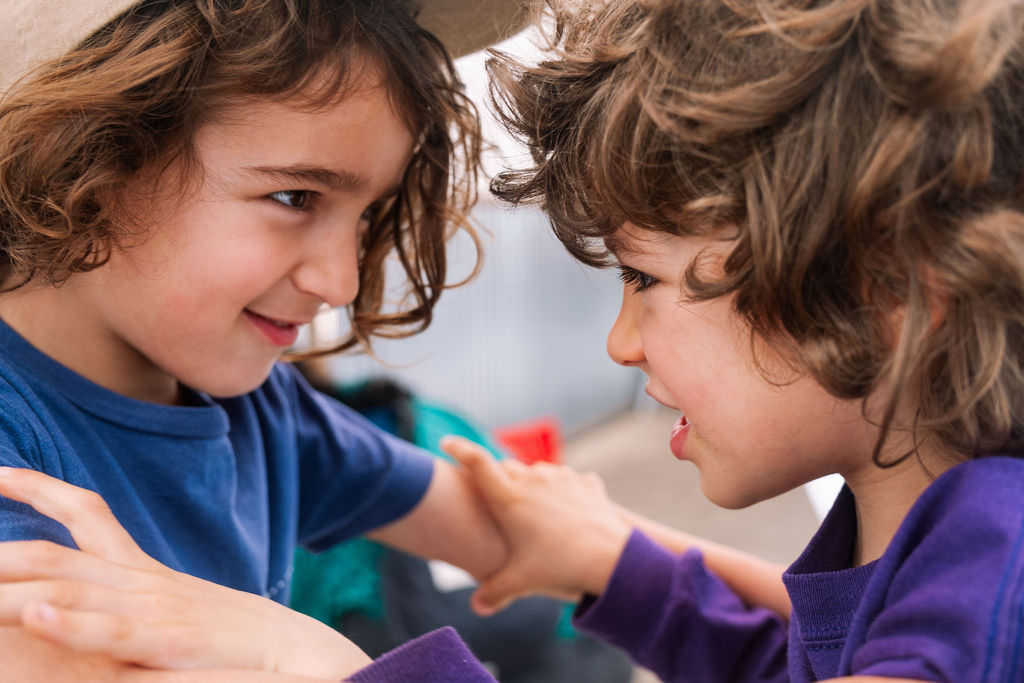Two children with curly hair are smiling as they touch each others shoulders, capturing a moment of kids friendship and joy. Dressed in casual clothes—one in blue, the other in purple—they appear to be playfully interacting during a lively photo session, set against a blurred background.