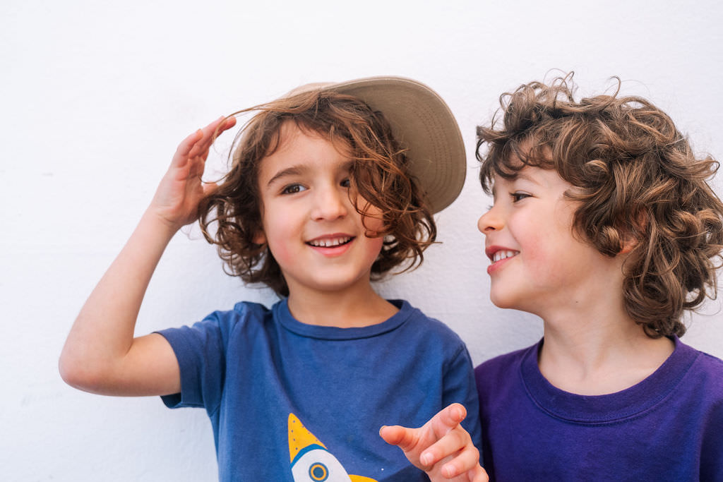 Two young children with curly hair smile and interact joyfully in a playful photo session against a plain white background. One child wears a blue shirt and a hat tilted sideways, while the other, in a purple shirt, looks at their friend, celebrating the spirit of kids friendship.