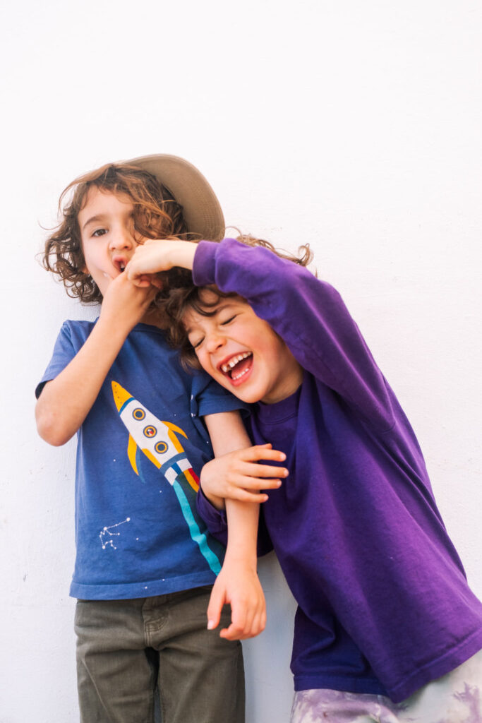 Two young children joyfully bond during a playful photo session against a white background. One wears a blue shirt with a rocket design and a hat, while the other, in a purple shirt, laughs with closed eyes. Their expressions of kids friendship are truly heartwarming.