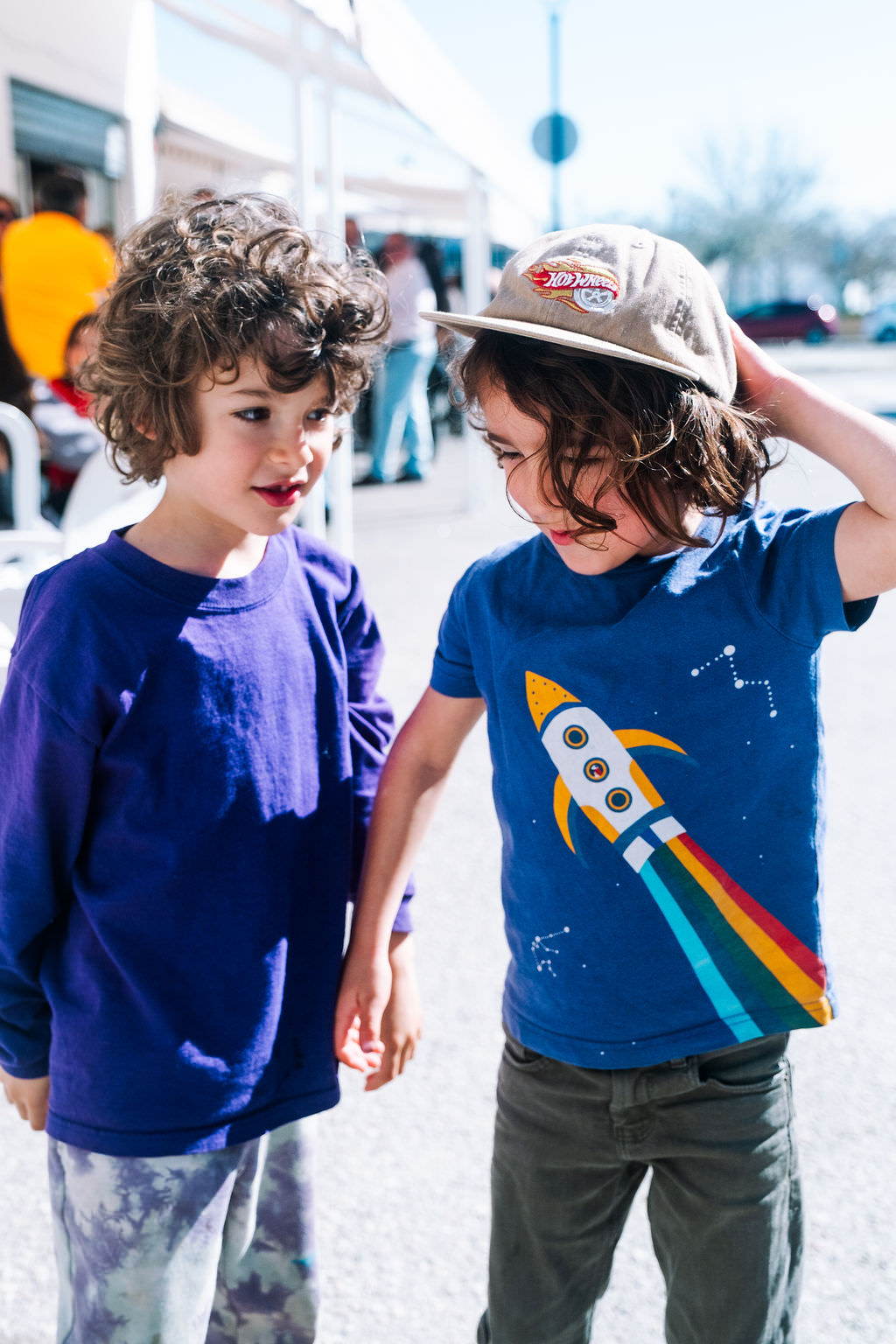 Two children stand outside, celebrating their friendship with playful smiles. One wears a blue shirt with a rocket design and a cap, while the other sports a purple shirt. They bathe in sunlight on the pavement, framed by a white building in this joyful photo session.
