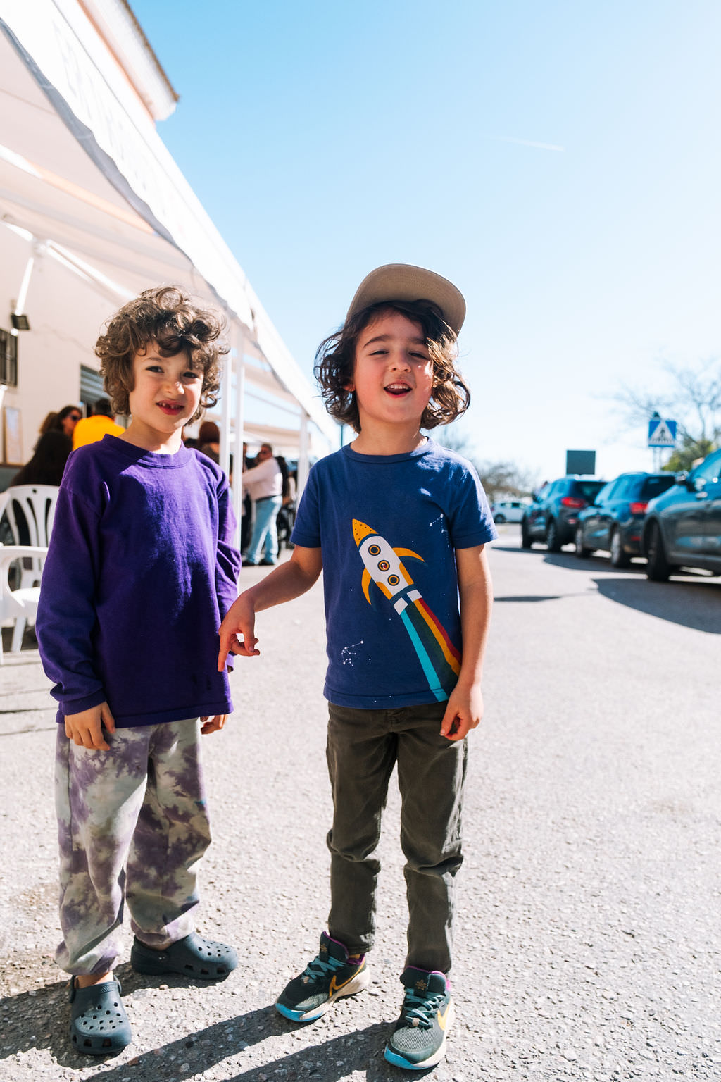 Two children stand on a sunny street, embodying playful friendship. The child on the left rocks a purple sweater and tie-dye pants, while the one on the right sports a blue rocket shirt and cap. Its as if they are in an impromptu photo session, with cars parked nearby and people gathering under a white canopy.