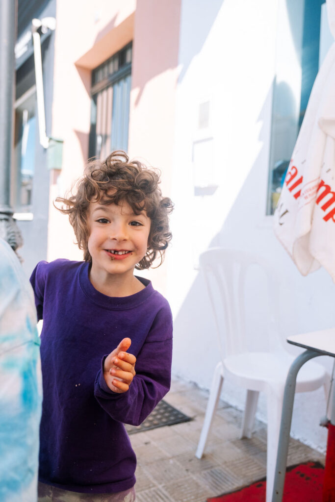 A child with curly hair, wearing a purple long-sleeve shirt, smiles playfully at the camera during a fun and playful photo session. The background includes a white plastic chair, a table, and parts of a building with light-colored walls.