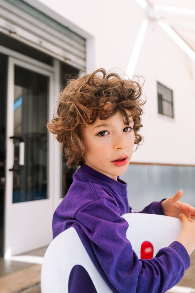 A young child with curly brown hair sits on a white chair outdoors, wearing a purple sweater. The patio setting, bathed in sunlight with a partially open door, hints at a playful photo session. The child gazes thoughtfully at the camera, capturing the essence of kids friendship.