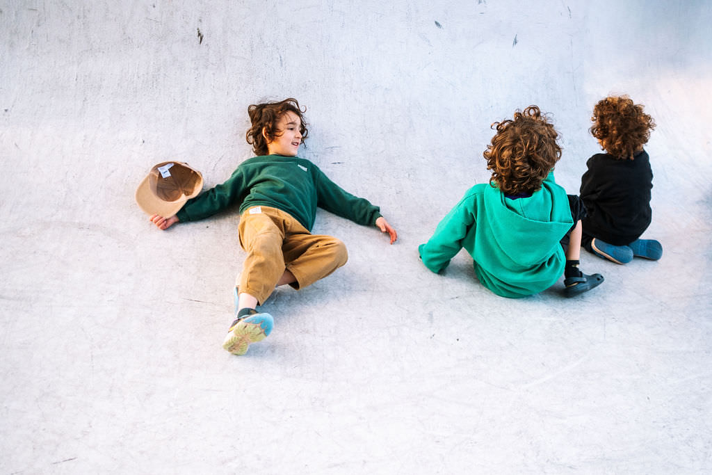 Three children with curly hair enjoy a playful photo session on a light-colored concrete surface. One child in a green hoodie lies down, while the others, dressed in black and turquoise, sit nearby. A tan cap rests on the ground, capturing a moment of kids friendship.