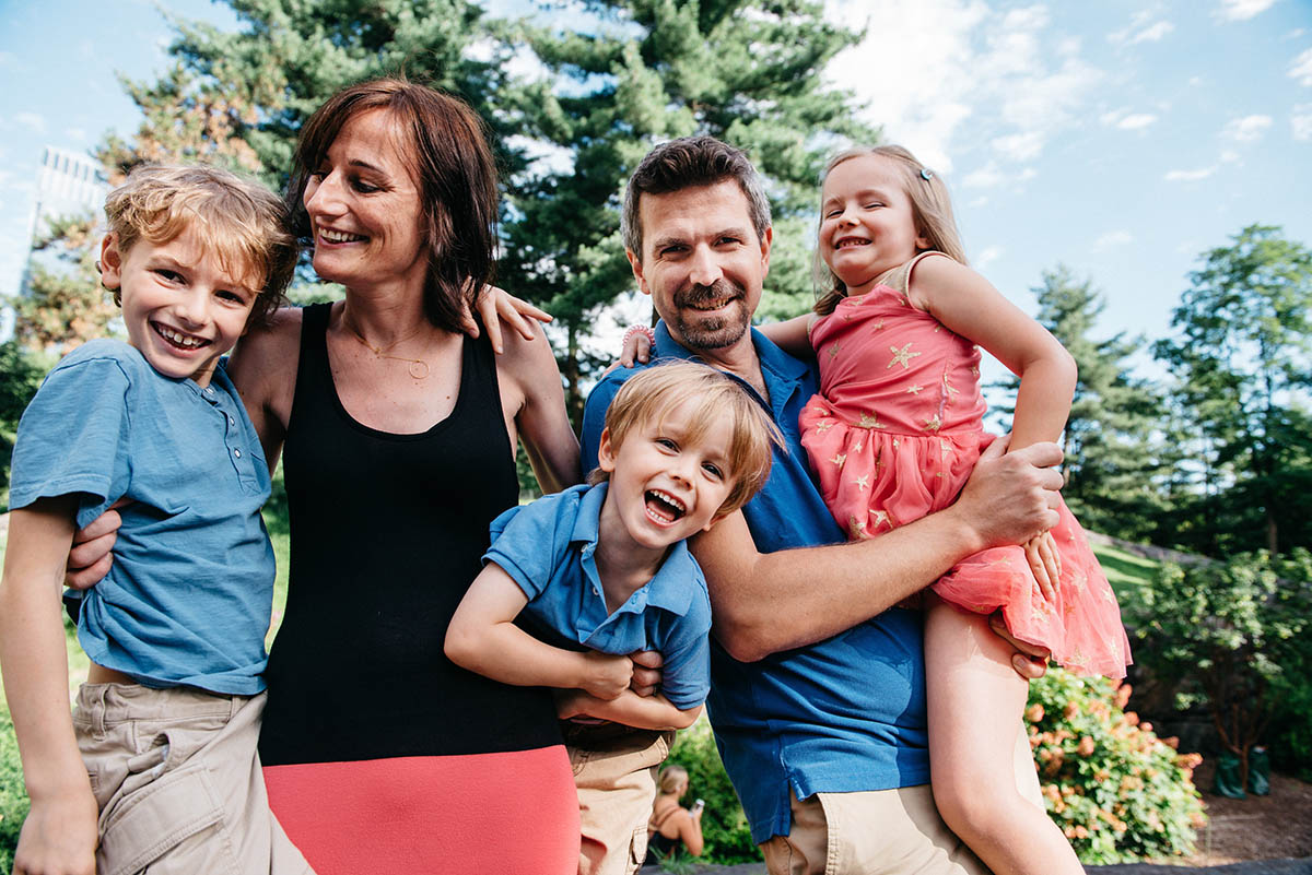 A smiling family of five poses outdoors on a sunny day, with trees in the background. Captured by an NYC family photographer, the father holds a young girl, while the mother and two boys stand close, all looking happy and cheerful.