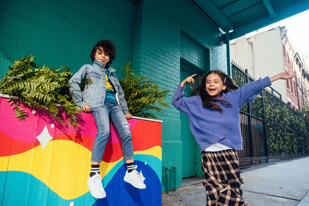 Two kids pose playfully outside on a city sidewalk. Captured by an NYC family photographer, one sits on a rainbow-painted planter in a denim jacket while the other, in a blue sweater and plaid skirt, smiles with arms raised amid green plants and buildings.