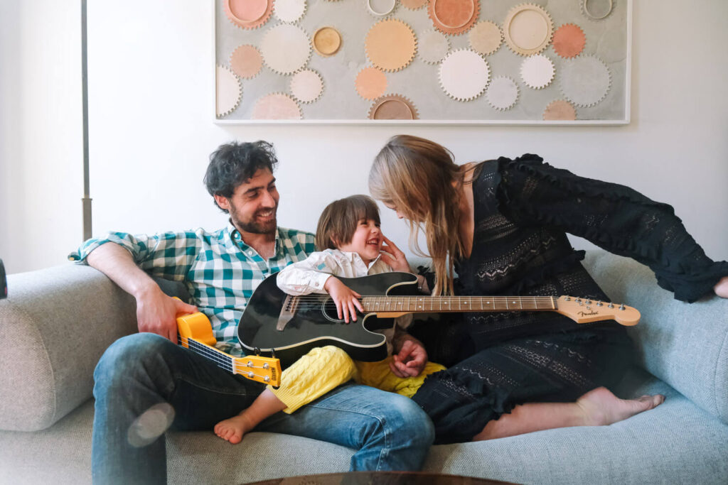 A man, woman, and child sit on a couch together, smiling and laughing. The man and woman each hold electric guitars as the child sits between them. Circular artwork hangs on the wall—capturing a joyful moment reminiscent of NYC Maternity Photography.