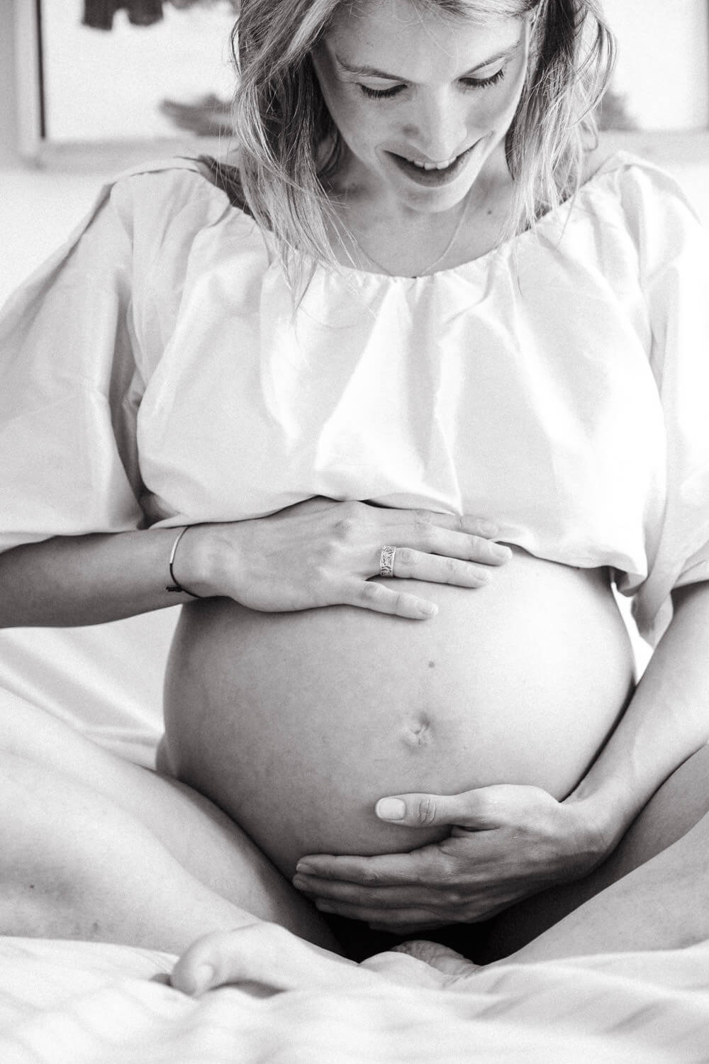 Black and white photo of a pregnant woman sitting cross-legged, smiling down as she gently cradles her bare baby bump. Captured in a candid NYC Maternity Photography style, she wears a loose, light-colored top.