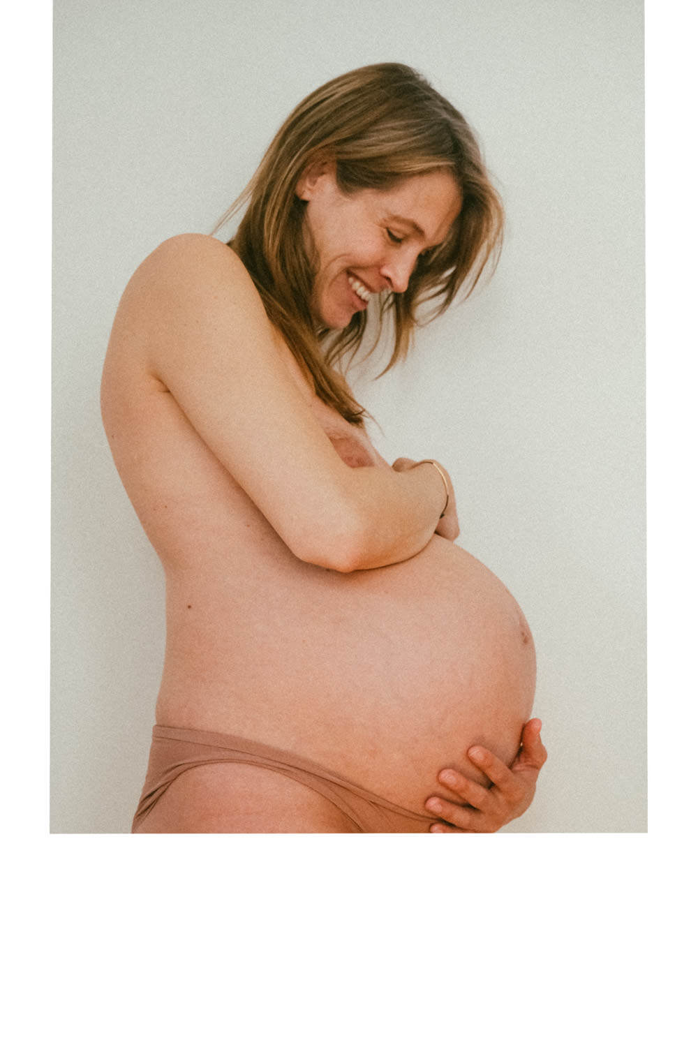 A smiling pregnant woman stands against a plain background, holding her bare belly with one hand and covering her chest with the other. She appears joyful and serene, embodying the radiant beauty often captured in NYC Maternity Photography.