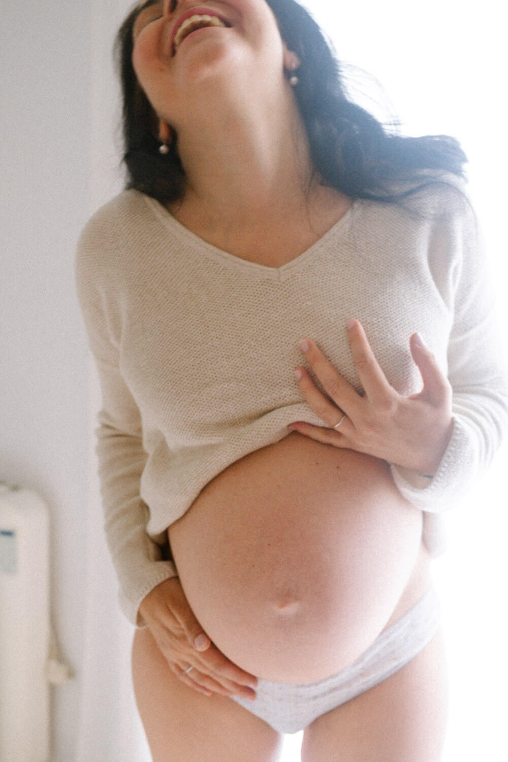 A pregnant woman smiles and tilts her head back, lifting her sweater to reveal her bare belly. She stands in soft, natural light—an intimate moment perfectly captured through NYC Maternity Photography.