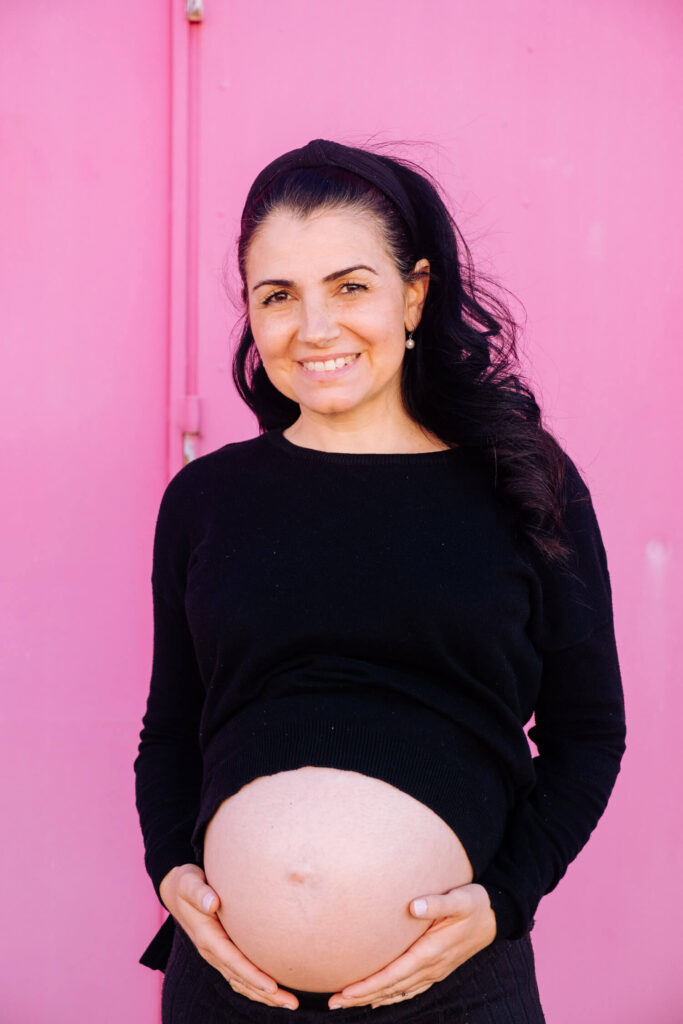 A smiling mom-to-be with long dark hair, wearing a black sweater, stands in front of a bright pink background and holds her bare baby bump during a maternity photoshoot.