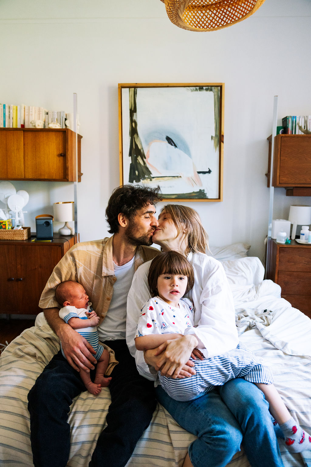 A couple sits on a bed, sharing a kiss as their NYC family photographer captures the moment. The man holds a baby, while the woman cradles a young child amidst cozy decor, wooden shelves, and a painting on the wall behind them.